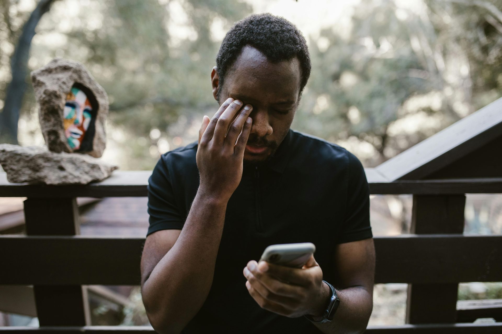 A man sits on a bench wiping his eye while holding a phone, looking distressed