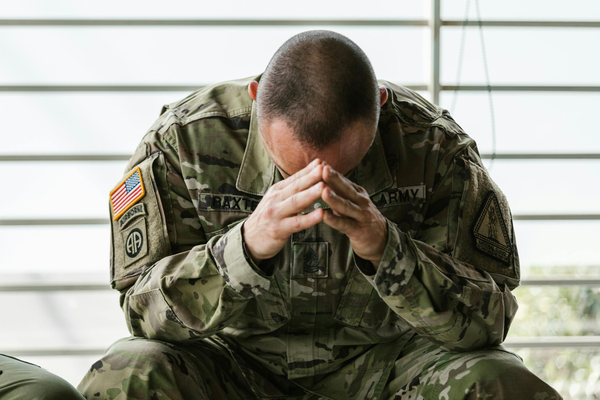 A soldier in camouflage uniform sits with his head bowed and hands clasped