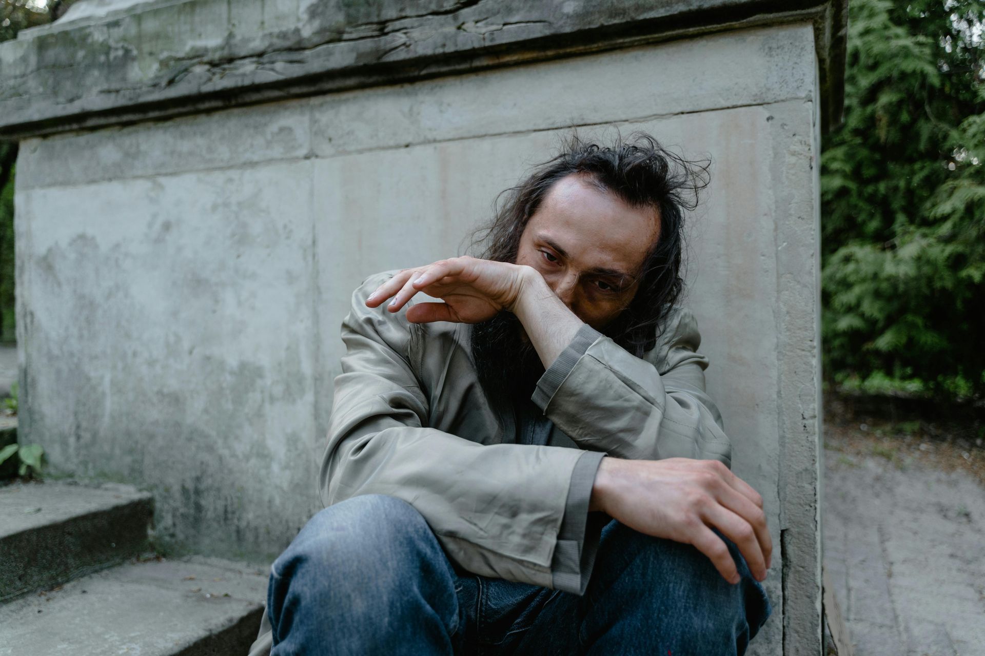 A man crouches against a concrete wall, hand to his face, looking uncertain