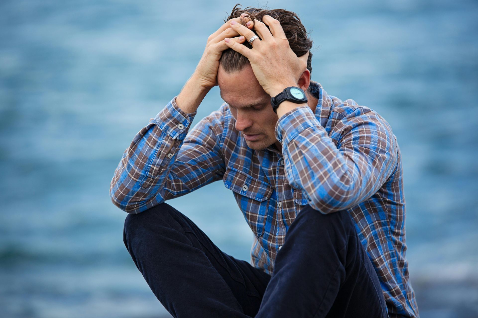 A man in a plaid shirt grips his head by the water, appearing anxious